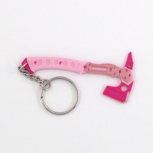May include: A pink and light pink miniature axe keyring with a silver key ring. The axe head and handle feature a geometric design. The keyring is a novelty item.