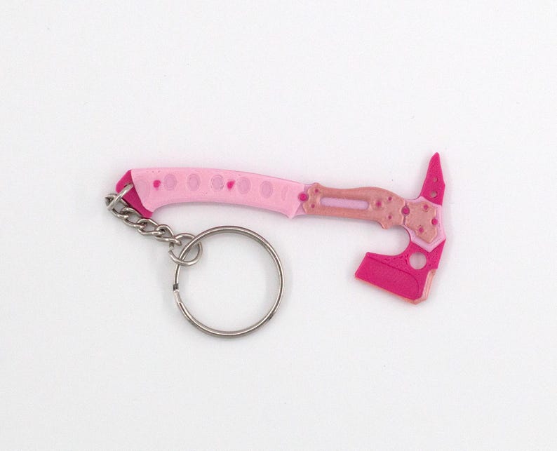 May include: A pink and light pink miniature axe keyring with a silver key ring. The axe head and handle feature a geometric design. The keyring is a novelty item.