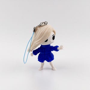 May include: A small figurine of a cartoon character with long, light-colored hair and a blue dress. The figure has a silver-colored clasp and a light blue string attached to the top of its head. The figurine is standing with its arms outstretched.