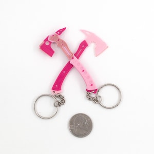 May include: Two pink and rose-coloured axe keychains crossed over each other, with silver key rings and chains. A coin is placed below the keychains. The axes have a detailed design and are made of a solid material.