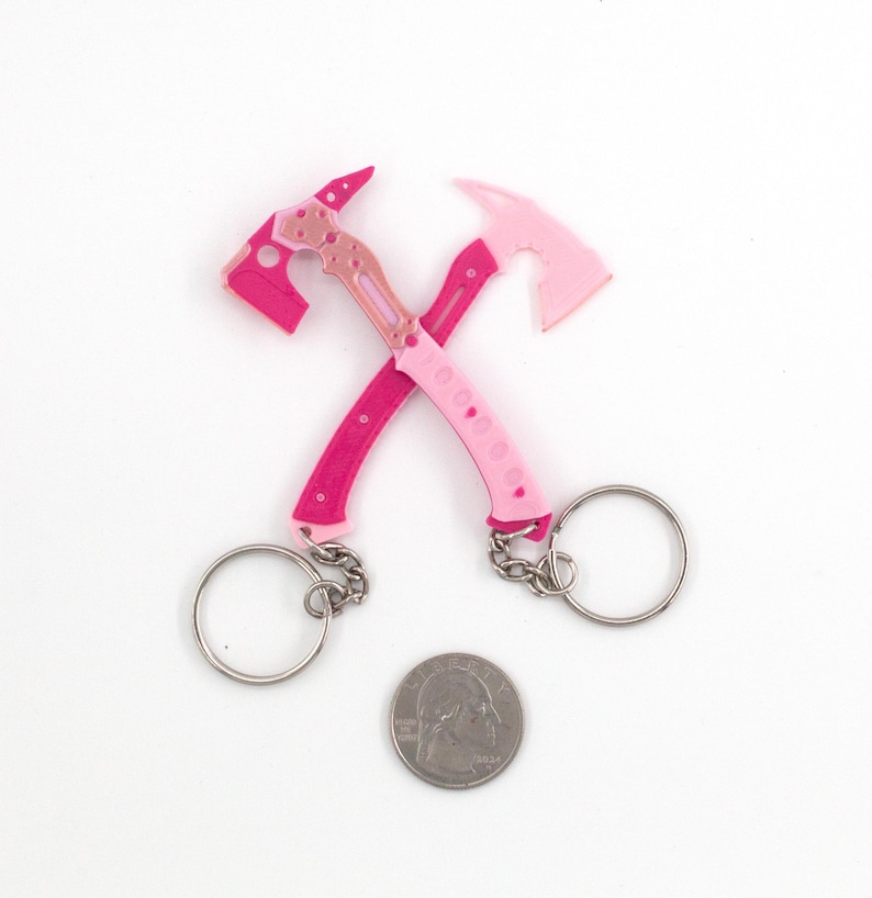 May include: Two pink and rose-coloured axe keychains crossed over each other, with silver key rings and chains. A coin is placed below the keychains. The axes have a detailed design and are made of a solid material.
