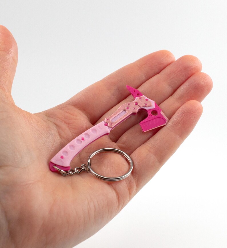 May include: A miniature pink and light pink axe keychain. The axe head is a bright pink, and the handle is light pink with small holes. A silver key ring is attached. The axe is a novelty item.