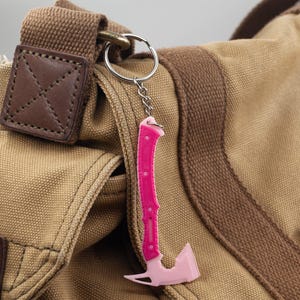 May include: A pink axe-shaped keyring with a silver chain and ring. The keyring is attached to a tan canvas bag with brown detailing and a leather patch.