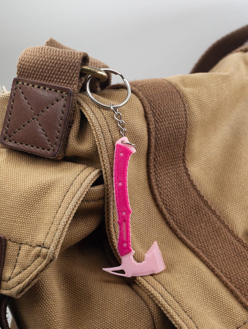 May include: A pink axe-shaped keyring with a silver chain and ring. The keyring is attached to a tan canvas bag with brown detailing and a leather patch.