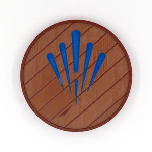 May include: A round, brown object with a wood-grain texture and a dark brown border. Five blue, stylized flame shapes radiate from the center. The design is reminiscent of a shield or emblem.