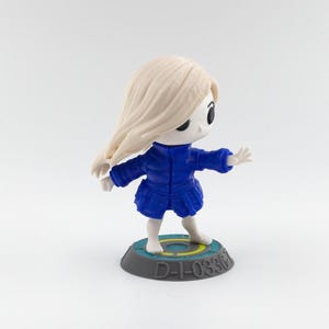 May include: A figurine of a character with flowing blonde hair and a blue outfit. The figure stands on a circular base with the text "D-1-0336". The figure has a stylized design with large black eyes.