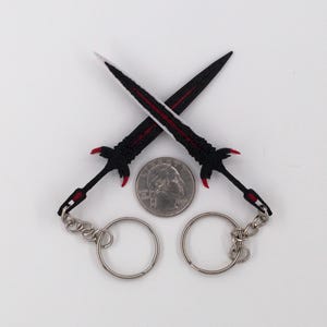 May include: Two black and red sword keychains crossed over a quarter. The swords have silver key rings and red accents on the hilt and blade. The blades are black with a red stripe down the center.