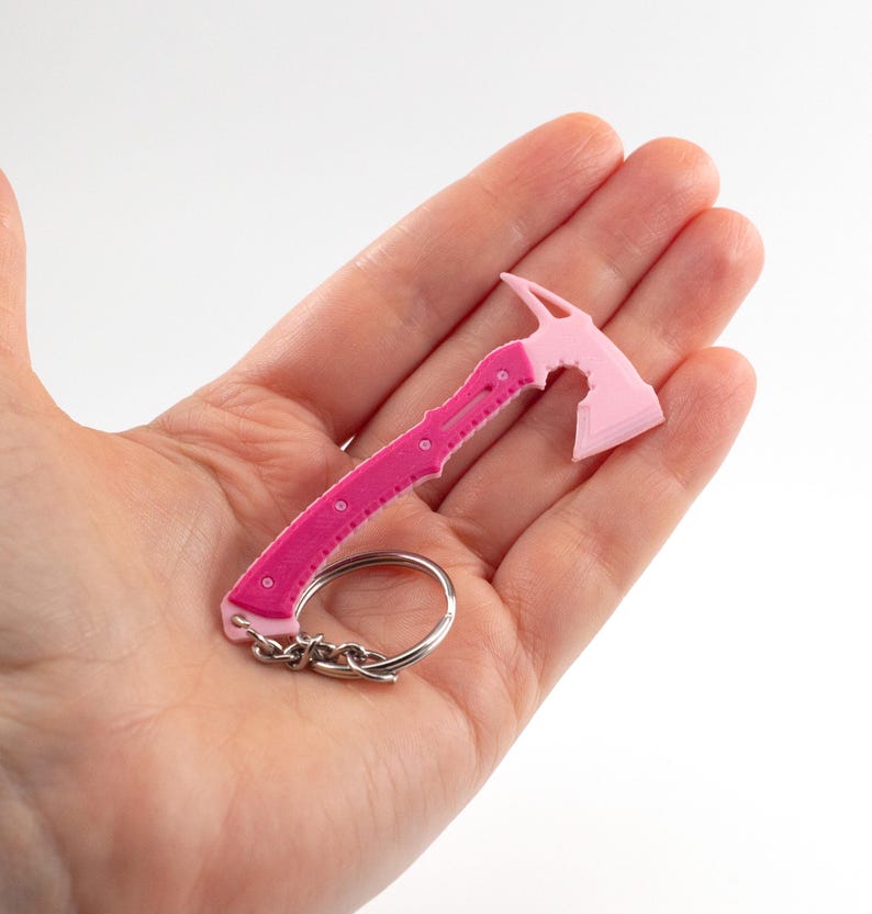 May include: A pink and magenta miniature axe keyring. The axe head is light pink, and the handle is magenta with circular details. A silver keyring and chain are attached.