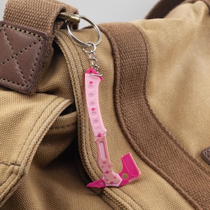 May include: A pink and white hatchet-shaped keyring, with floral detailing, hanging from a silver ring. The keyring is attached to a brown canvas bag with a leather patch and brown strap.