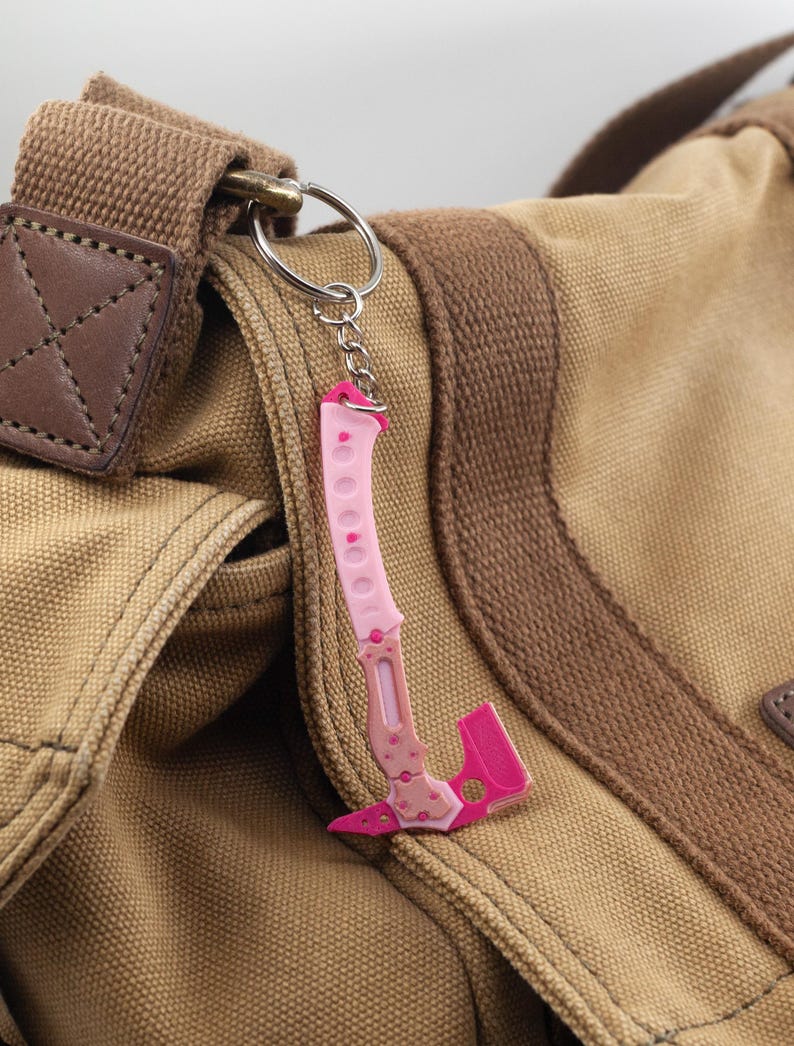 May include: A pink and white hatchet-shaped keyring, with floral detailing, hanging from a silver ring. The keyring is attached to a brown canvas bag with a leather patch and brown strap.