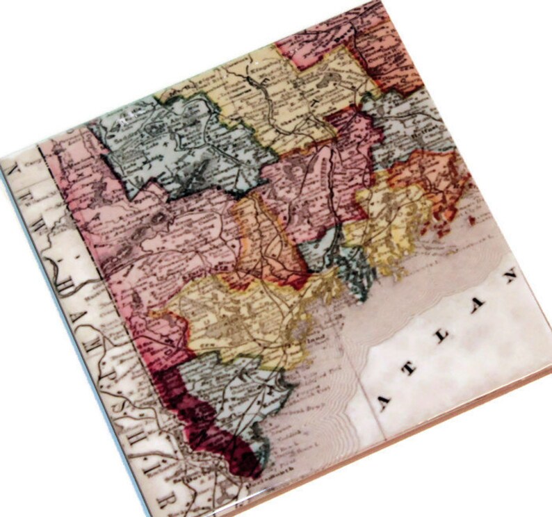 May include: An antique map of New England, printed on a ceramic coaster. The map shows the states of New Hampshire, Vermont, Massachusetts, Rhode Island, and Connecticut. The map is in shades of pink, yellow, green, and blue.