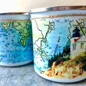May include: Two white enamel mugs with a nautical theme. One mug features a map with the text "Bass Harbor Head Light" and the other mug features a lighthouse with the text "Cranberry Isle" and "Western Way".