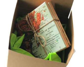 May include: A stack of map-themed coasters tied with twine, inside a brown cardboard box with green tissue paper. The coasters feature a vintage map of Boston, Massachusetts, with orange and green hues. The box is open, revealing the coasters.