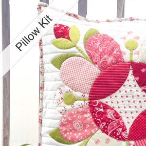 May include: A square pillow kit featuring a quilted white background with a large floral applique design. The flower petals and leaves are crafted from various red, pink, and green fabrics with floral, paisley, and gingham patterns. Text reads Pillow Kit.