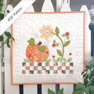 May include: A quilted wall hanging featuring a pumpkin and floral design. The pumpkin is orange with green leaves, and the flowers are yellow and orange. The background is white with a floral border. The text "PDF Pattern" is in the upper left corner.