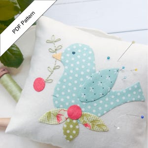 Könnte beinhalten: Ein weißes Stoff-Nadelkissen mit einem Applikation eines blau gepunkteten Vogels. Der Vogel hält einen Zweig mit einer roten Kirsche. Mehrere Nadeln sind eingesteckt. Der Text "PDF Pattern" befindet sich oben links.