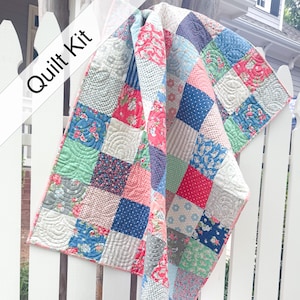 May include: A patchwork quilt kit featuring a variety of square fabric blocks in shades of red, blue, green, and white with floral, polka dot, and striped patterns. The quilt has a pink border and visible curved machine quilting stitches.