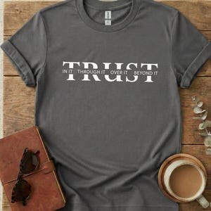 Könnte beinhalten: Ein graues T-Shirt mit dem Wort "TRUST" in weißen Blockbuchstaben. Unter dem Wort "TRUST" stehen die Worte "IN IT THROUGH IT OVER IT BEYOND IT". Das Shirt ist an den Ärmeln gefaltet und liegt auf einer Holzoberfläche.