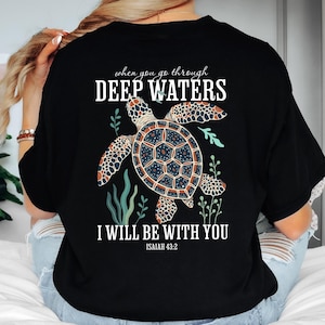 Könnte beinhalten: Schwarzes T-Shirt mit einem Schildkröten-Design und dem Text "when you go through DEEP WATERS I WILL BE WITH YOU ISAIAH 43:2". Die Schildkröte hat einen gemusterten Panzer und ist von Wasserpflanzen umgeben.
