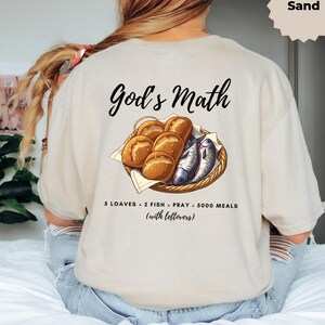 Könnte beinhalten: Beigefarbenes T-Shirt mit dem Text "God's Math" und einer Illustration von Brot und Fisch in einem Korb. Darunter steht "5 Brote, 2 Fische, Gebet, 5000 Mahlzeiten (mit Resten)".