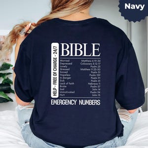 Könnte beinhalten: Marineblaues T-Shirt mit weißem Text. Das Wort "BIBLE" steht oben, gefolgt von einer Liste emotionaler Zustände und zugehörigen Bibelversen. Das Shirt enthält auch den Text "HELP - FREE OF CHARGE - 24/7" und "EMERGENCY NUMBERS".