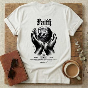Puede incluir: Camiseta blanca con la palabra "Faith" en fuente gótica, sobre un gráfico de manos sosteniendo un globo. Debajo, las palabras "ESTD 2026" y un versículo bíblico. La camiseta está sobre una superficie de madera.