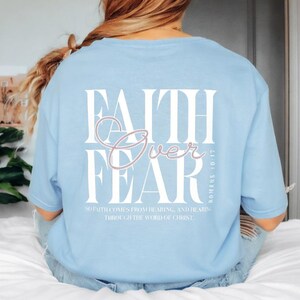 Könnte beinhalten: Hellblaues T-Shirt mit den Worten "FAITH Over FEAR" in weißer und rosa Schrift. Der Text "Romans 10:17" ist darunter gedruckt, mit einem Zitat über den Glauben. Das T-Shirt hat einen lässigen Stil.
