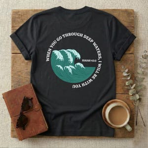Könnte beinhalten: Ein schwarzes Kurzarm-T-Shirt mit einer kreisförmigen Grafik aus türkisfarbenen Meereswellen. Der Text um die Wellen herum lautet "WHEN YOU GO THROUGH DEEP WATERS, I WILL BE WITH YOU" und "ISAIAH 43:2" in weißen Großbuchstaben.