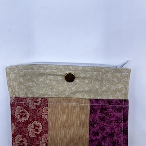 May include: A handmade fabric wallet with a beige floral pattern top and a color block design on the bottom. The bottom features a burgundy floral print, a tan section, and a purple floral print. The wallet has a zipper closure and a brass snap.