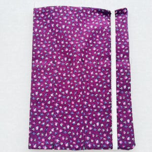 May include: A rectangular, folded fabric item with a deep purple floral pattern. The pattern features small white, pink, and blue flowers. A white, lace-trimmed ribbon runs along one edge.