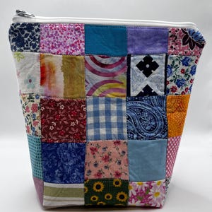 May include: A patchwork zippered pouch with a variety of fabric squares in floral, paisley, and geometric patterns. The pouch features a white zipper and a light-colored lining. The overall design is a mix of colors and textures.