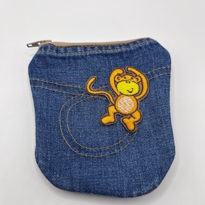 May include: A small denim pouch featuring a dark blue fabric with visible orange stitching details. An embroidered patch of a yellow and brown monkey is centered on the front. The pouch includes a tan zipper closure at the top edge.