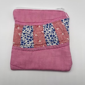 May include: A small rectangular fabric pouch featuring a solid pink top and bottom section. The center panel consists of alternating vertical strips of red and white checkered fabric and blue and white floral patterned fabric with a zipper.