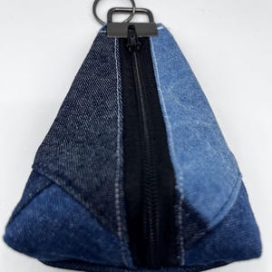May include: A denim triangle-shaped pouch with a black zipper and a metal key ring. The pouch is constructed from various shades of blue denim, creating a patchwork effect. The zipper runs down the center, providing access to the interior.