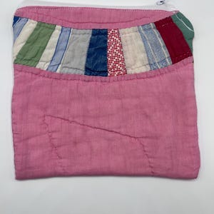 May include: A rectangular zippered pouch made of pink fabric with a quilted texture. The top section features a horizontal band of multicolored fabric strips in shades of green, blue, white, and red, stitched together with visible white thread.