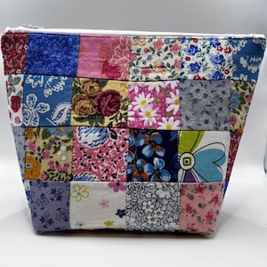 May include: A patchwork zippered pouch with a white zipper and lining. The pouch is made of various floral fabric squares in shades of blue, pink, yellow, and red. The bag has a rectangular base and tapers upwards.