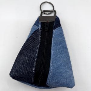 May include: A denim coin purse with a black zipper and a metal key ring. The purse is made of patchwork denim in shades of blue and navy. The zipper runs down the center of the purse.