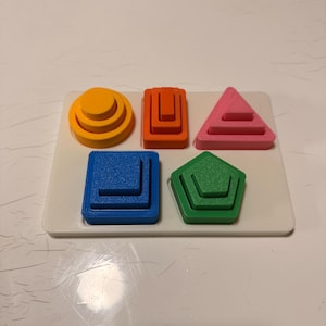 Puede incluir: Un rompecabezas de madera rectangular blanco con cinco formas geométricas de colores: un círculo escalonado amarillo, un bloque naranja en forma de U, un triángulo rosa, un bloque azul en forma de L y un pentágono verde.