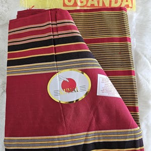 Op de afbeelding: Gevouwen textiel met een rode basis en horizontale strepen in zwart, geel en wit. De verpakking toont "PRIDE OF UGANDA". Een gouden sticker met tekst is ook zichtbaar.