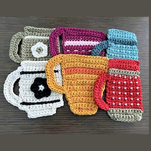 May include: A collection of handmade crochet mug rugs in various colors. The rugs are shaped like mugs and feature different designs, including floral accents and striped patterns. The color palette includes white, red, yellow, purple, and blue.