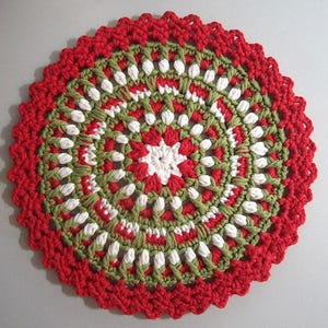 May include: A round, crocheted doily in red, green, and white. The outer edge is a scalloped red, with a pattern of green and white stitches forming a circular design. The center features a white starburst.