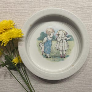 Könnte beinhalten: Eine weiße Keramikschale mit erhöhtem Rand. Das Innere der Schale zeigt eine Vintage-Illustration von drei Kindern auf einem Feld. Gelbe Blumen mit grünen Stielen befinden sich in der unteren linken Ecke.