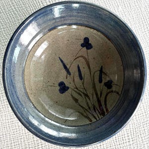 Può includere: Una ciotola rotonda in ceramica con interno beige maculato e un ampio bordo blu lucido. Il centro della ciotola presenta un disegno botanico dipinto a mano con fiori blu scuro e steli verdi che contrastano con la base chiara maculata.