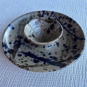 May include: A ceramic serving dish featuring a central raised bowl surrounded by a wide, flat rim. The piece has a speckled beige base color with dark blue and brown paint splatters across the entire surface, creating a textured appearance.