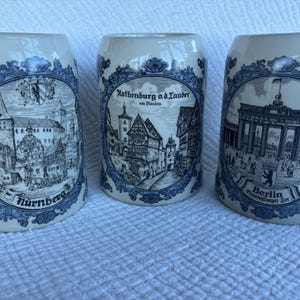 May include: Three off-white ceramic beer steins featuring blue decorative borders and black and white illustrations of German landmarks. The mugs are labelled with the text Nurnberg, Rothenburg o.d. Tauber, and Berlin Brandenburger Tor.