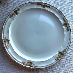 Puede incluir: Una fuente de cerámica blanca con borde festoneado y ribete dorado. La fuente presenta detalles florales dorados alrededor del borde. El plato es redondo y parece sin usar.