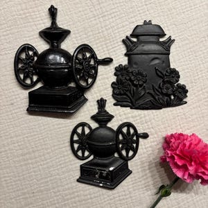 May include: Two black cast iron wall plaques shaped like vintage coffee grinders with side wheels and a central black cast iron plaque featuring a milk can design with embossed floral details, accompanied by a single bright pink carnation flower.