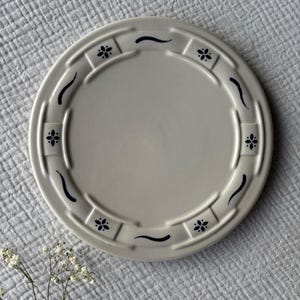 May include: A round, off-white ceramic plate with a decorative border. The border features a repeating pattern of dark blue floral designs and curved lines. The plate is set on a light gray, textured surface.