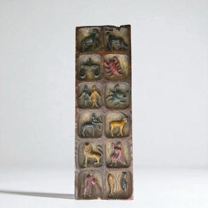 May include: A rectangular, vertical sculpture with twelve carved panels, each depicting a zodiac sign. The signs are rendered in relief and painted with muted colors, including green, red, and gold. The overall aesthetic is rustic and antique.