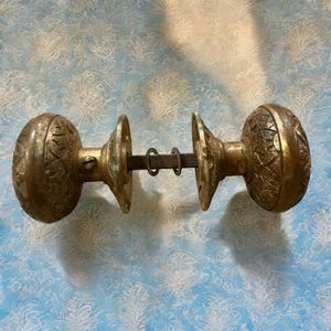 Könnte beinhalten: Antikes Türknauf-Set aus Messing. Das Set besteht aus zwei verzierten, runden Knöpfen mit detaillierten floralen Mustern, die durch eine Metallstange und Montageplatten verbunden sind. Das Messing hat eine Vintage-Patina. Für eine Tür.